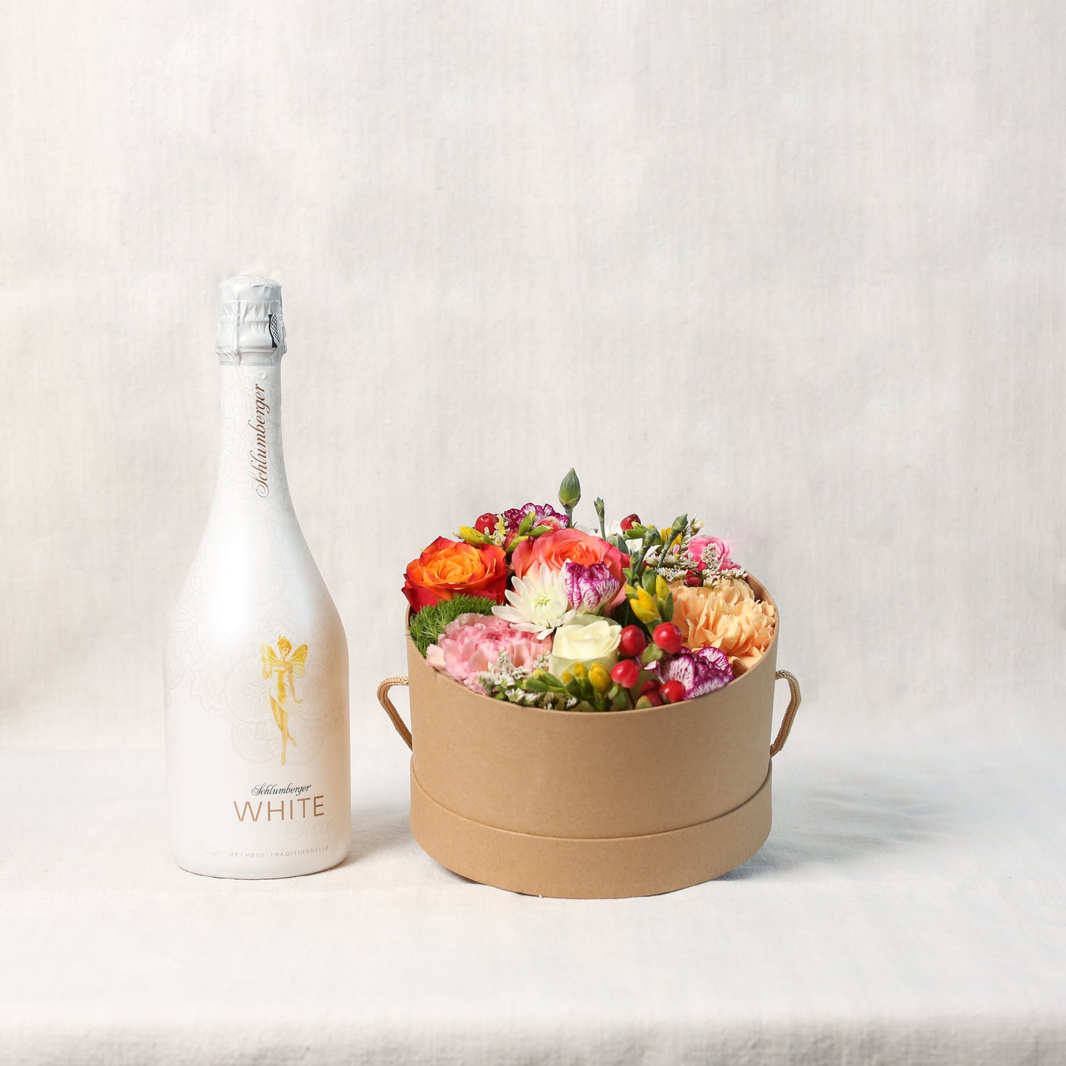 Seitansicht - Bunt gemischte Saisonblumen, gesteckt in einem naturfarbenen Rundkarton und dazu eine Flasche Schlumberger Sekt