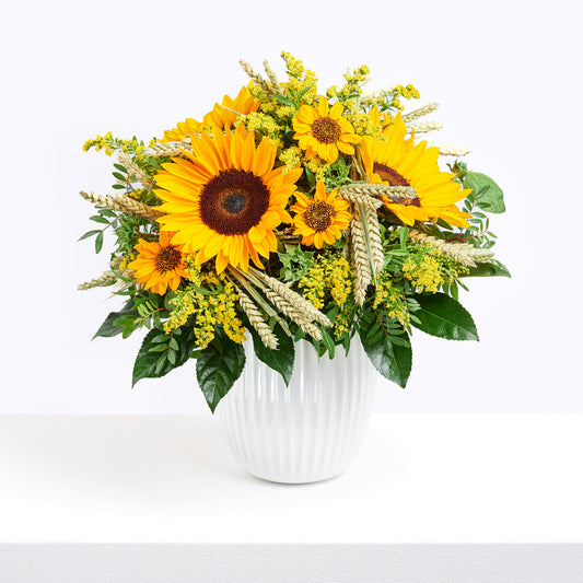 Strauß der Woche, in einer weißen Vase, mit Sonnenblumen, Getreideähren und weiteren kleinen Blumen in Gelbtönen. Dazu diverses Grün. Seitenansicht.