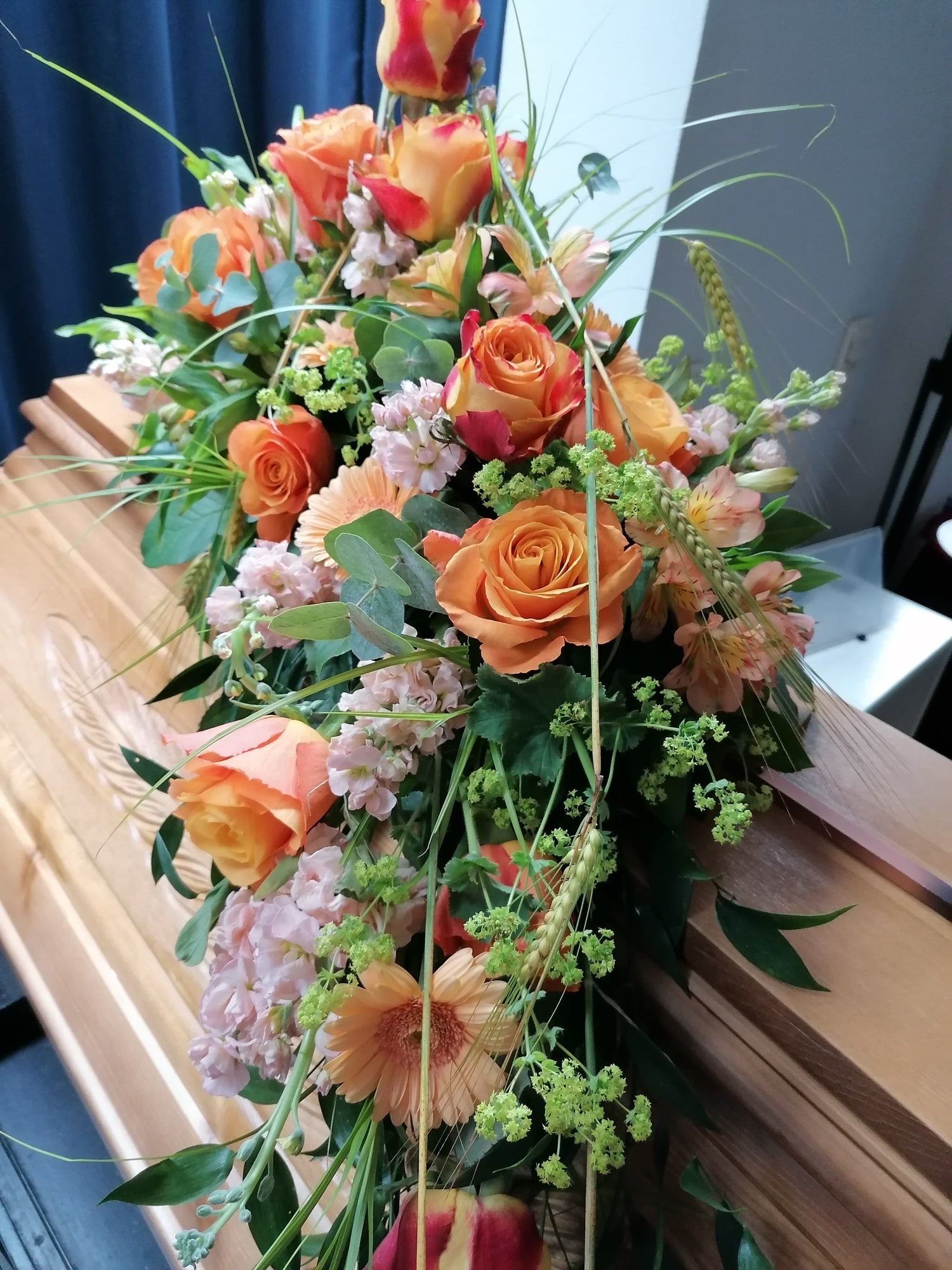 Längliches, abfließendes Sargbukett mit orangen Rosen und diversen anderen blassrosa Blumen mit grünen Beiwerk auf einem Sargdeckel
