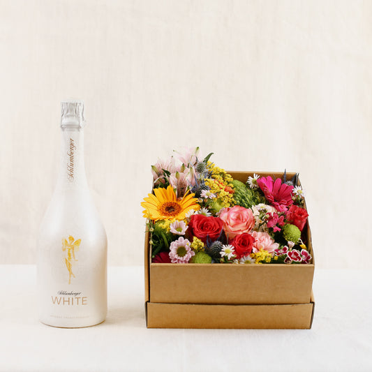 Eine Flasche Edler Schlumberger-Sekt, Sparkling Brut Klassik „ganz in weiß“ mit goldenem Engel auf weißem Grund mit einer mit bunten Blumen gefüllten Blumen-Box